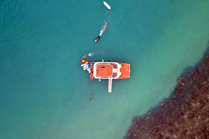 The catamaran anchored beside the reef edge, seen from above: shallow coral on one side, deeper sand on the other