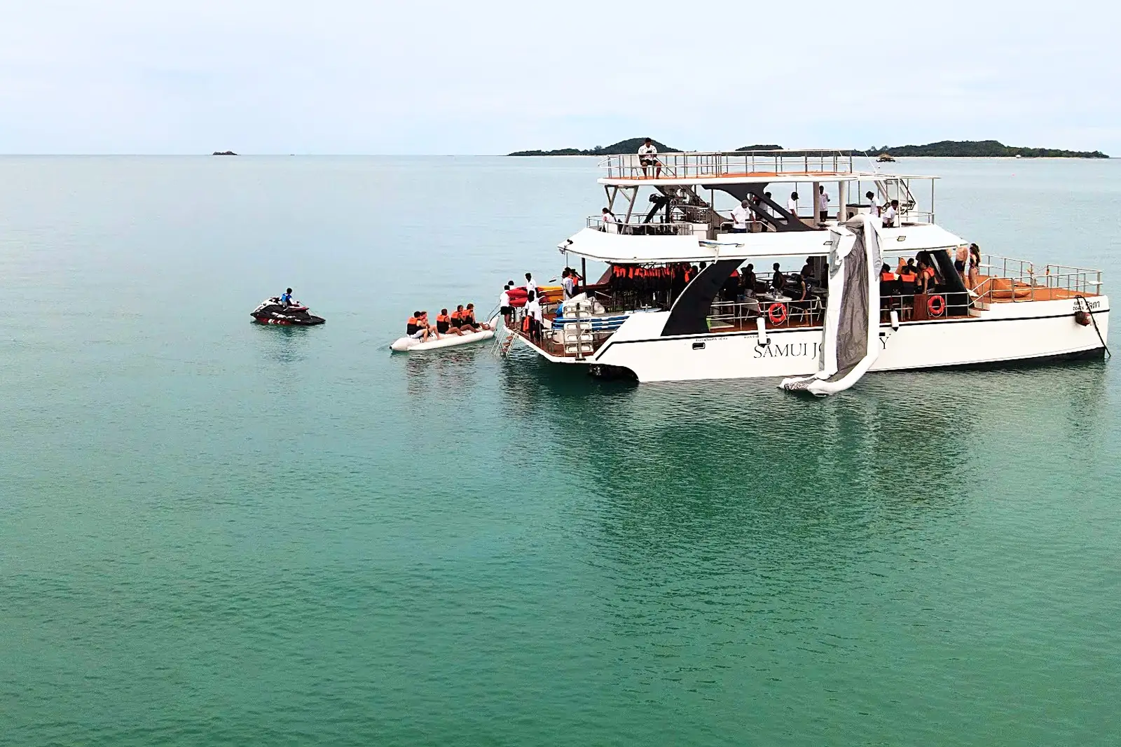 Guests in life jackets on the banana boat beside the catamaran, with the jet ski ready to tow