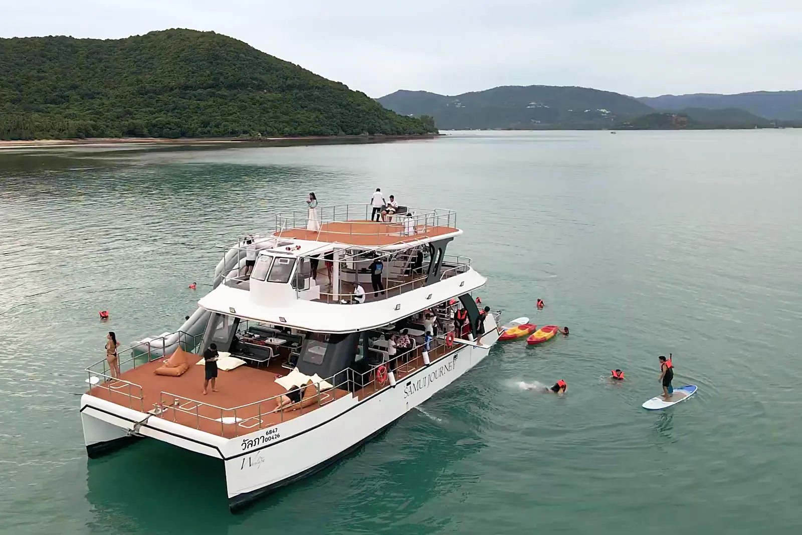 A guest stand-up paddleboarding beside the anchored catamaran, with green hills behind