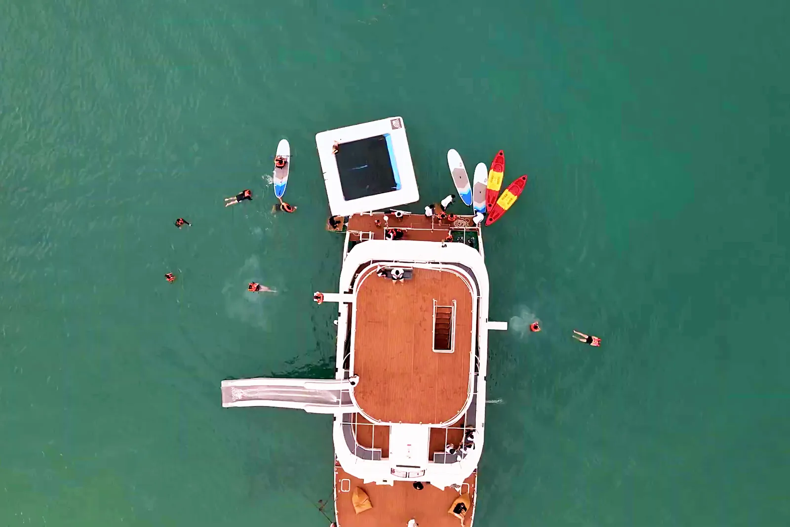 Top-down view of the floating sea pool, paddleboards and kayaks at the catamaran bow, with guests swimming around